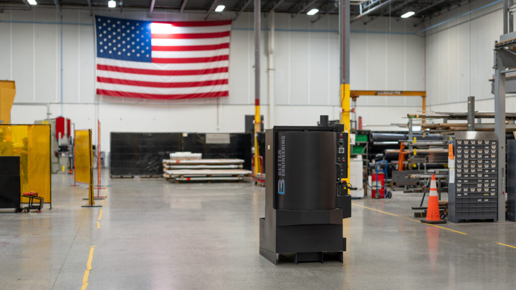 A black Better Engineering parts washer sits centered in a spacious manufacturing facility with an American flag hanging on the wall in the background.